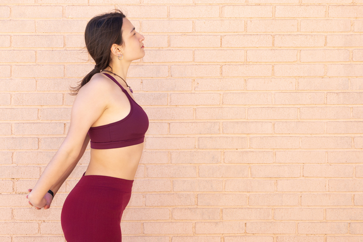 A young Caucasian woman dressed in a top and leggings is doing arm, chest and back stretches next to a brick wall.