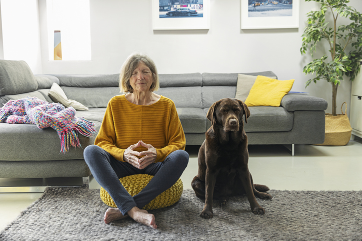 A senior woman (in her 70s who is undergoing targeted treatment for Stage 4 non small-cell lung cancer (NSCLC)) meditates in her living room with her chocolate labrador dog. Their faces displays their positivity.