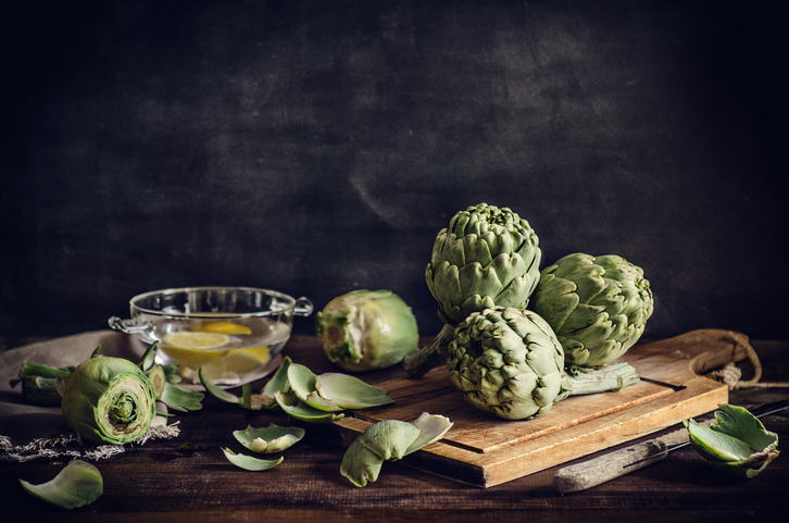 Raw artichokes on a wooden cutting board, with a bowl and some lemon slices, on a wooden surface and a slate background.